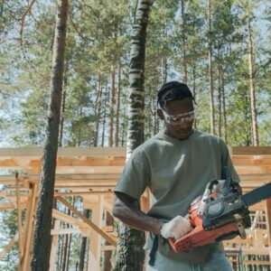 A man at a construction site in a forest, operating a chainsaw amidst tall trees.