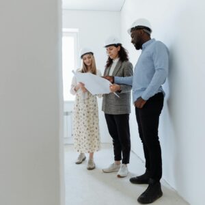 Three architects in hard hats review blueprints in a bright, empty white room.